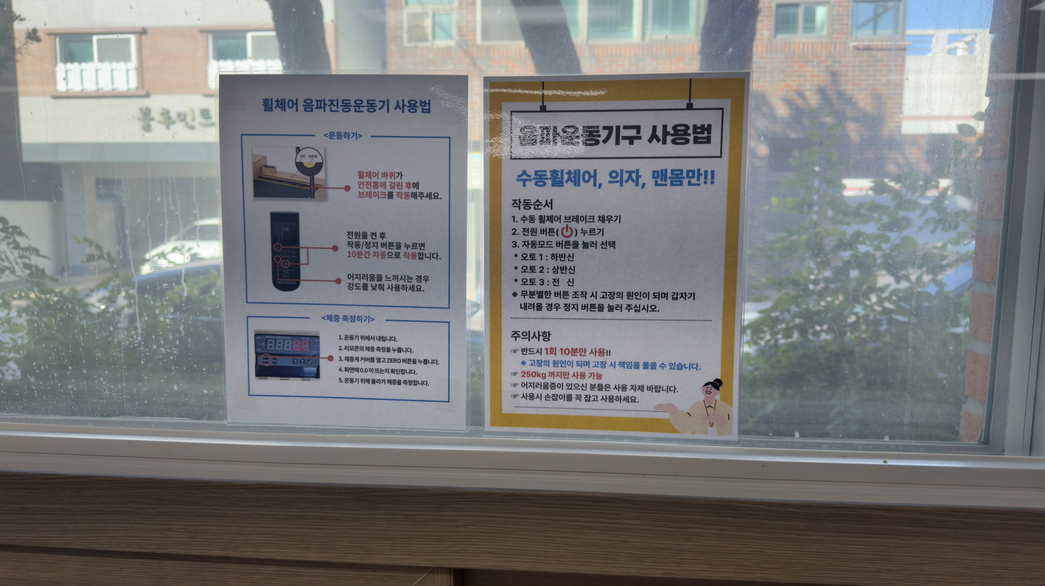 복지관 창문에 붙은 휠체어 음파진동운동기 사용 안내문 두 장. 왼쪽 안내문은 ‘휠체어 음파진동운동기 사용법’으로, 휠체어를 기기에 고정하고 전원을 켠 후 10분간 자동으로 작동한다는 설명과 어지럼증 시 강도를 낮추라는 주의사항이 적혀 있다. 오른쪽 안내문은 ‘음파운동기구 사용법’으로, 수동 휠체어·의자·맨몸만 사용 가능하며 작동 순서와 안전 주의사항(13분 이내 사용, 250kg까지 사용 가능, 손잡이 잡고 사용 권장 등)이 상세히 안내되어 있다.
