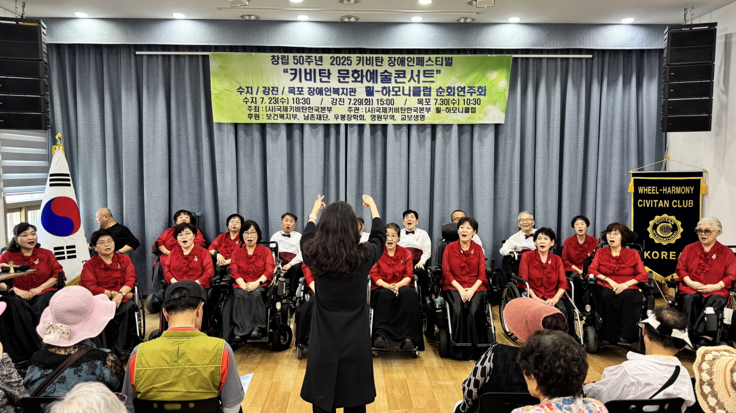 지휘자가 앞에 서서 손을 들어 합창을 이끄는 가운데, 휠-하모니클럽 단원들이 힘차게 노래를 부르는 장면