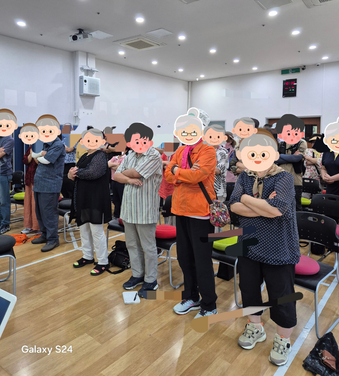 건강교육 참여자들이 자리에서 일어나 팔짱을 끼고 서 있으며, 강사의 안내에 따라 간단한 동작을 함께 하고 있습니다