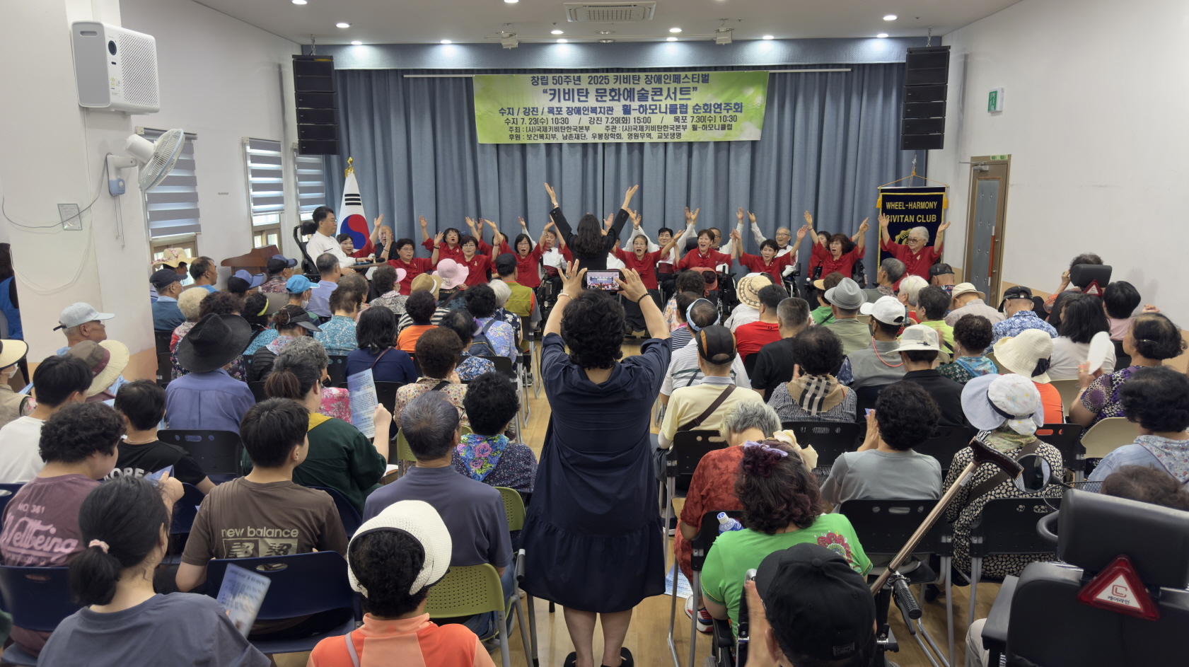 관객으로 가득 찬 강당에서, 장애인들이 함께 관람하는 가운데 휠-하모니클럽 단원들이 두 팔을 높이 들어 올리며 합창 공연을 펼치는 장면