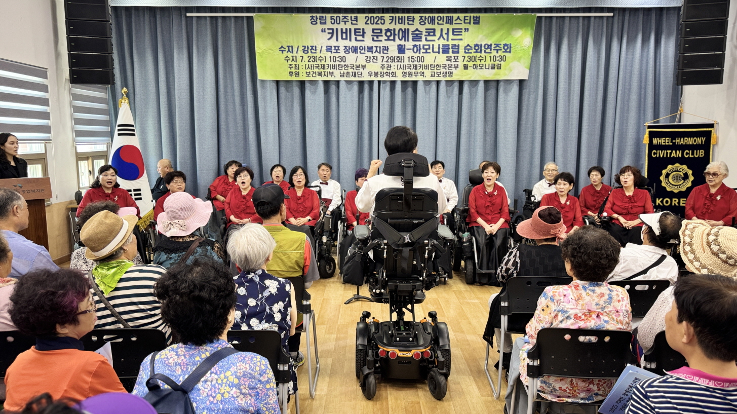 전동휠체어를 탄 휠-하모니클럽 단원이 무대 앞에서 지휘를 맡고, 단원들이 노래하며 관객인 장애인들이 집중해 공연을 보는 모습