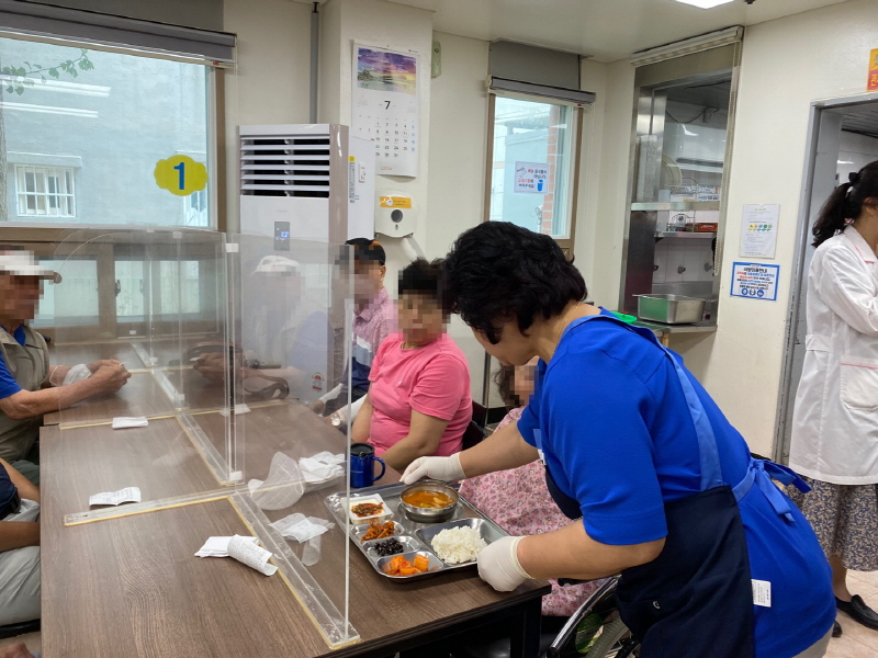 민주당 여성위원회 봉사자가 파란색 조끼를 입고 장애인에게 밥과 국이 담긴 식판을 나누어주는 모습