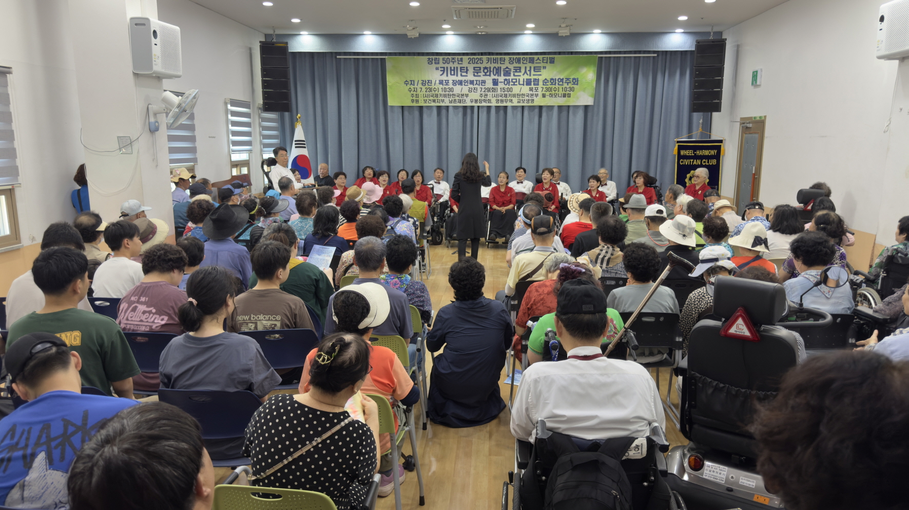 많은 장애인 관객이 객석을 메운 가운데, 휠-하모니클럽 단원들이 무대에서 지휘자의 손짓에 맞춰 노래하는 모습