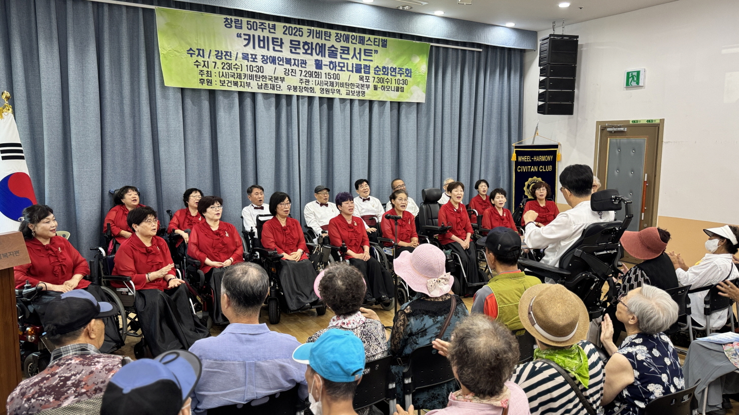 휠-하모니클럽 단원들이 붉은색 의상을 입고 무대 앞에 앉아 노래를 부르고 있으며, 객석의 장애인들이 공연을 관람하는 모습