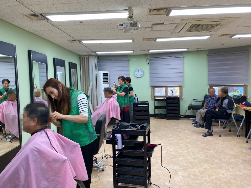 하당동새마을부녀회 봉사자들이 남성 이용자의 머리를 다듬고 있으며, 뒤쪽 의자에는 머리 손질을 기다리는 어르신들이 앉아 있는 모습