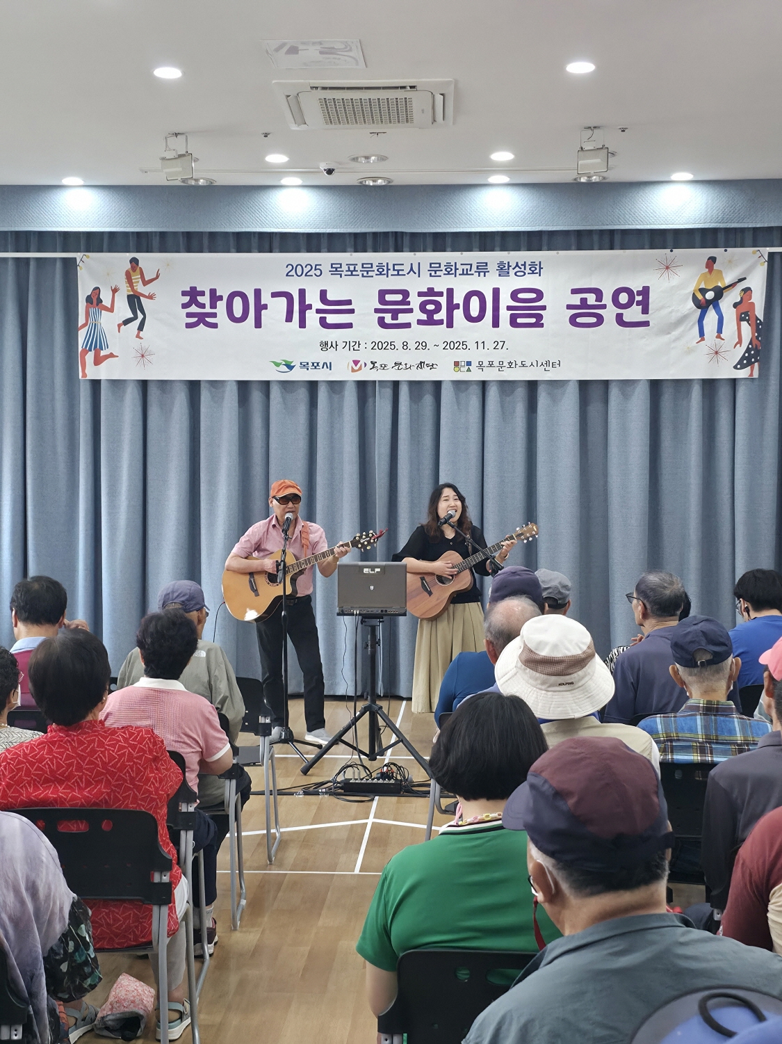 남성과 여성 2명의 듀오가 각각 기타를 연주하며 노래하고 있다. 앞줄부터 어르신 관객들이 빼곡히 앉아 무대를 바라보고 있으며, 무대 위에는 ‘찾아가는 문화이음 공연’ 현수막이 걸려 있다.