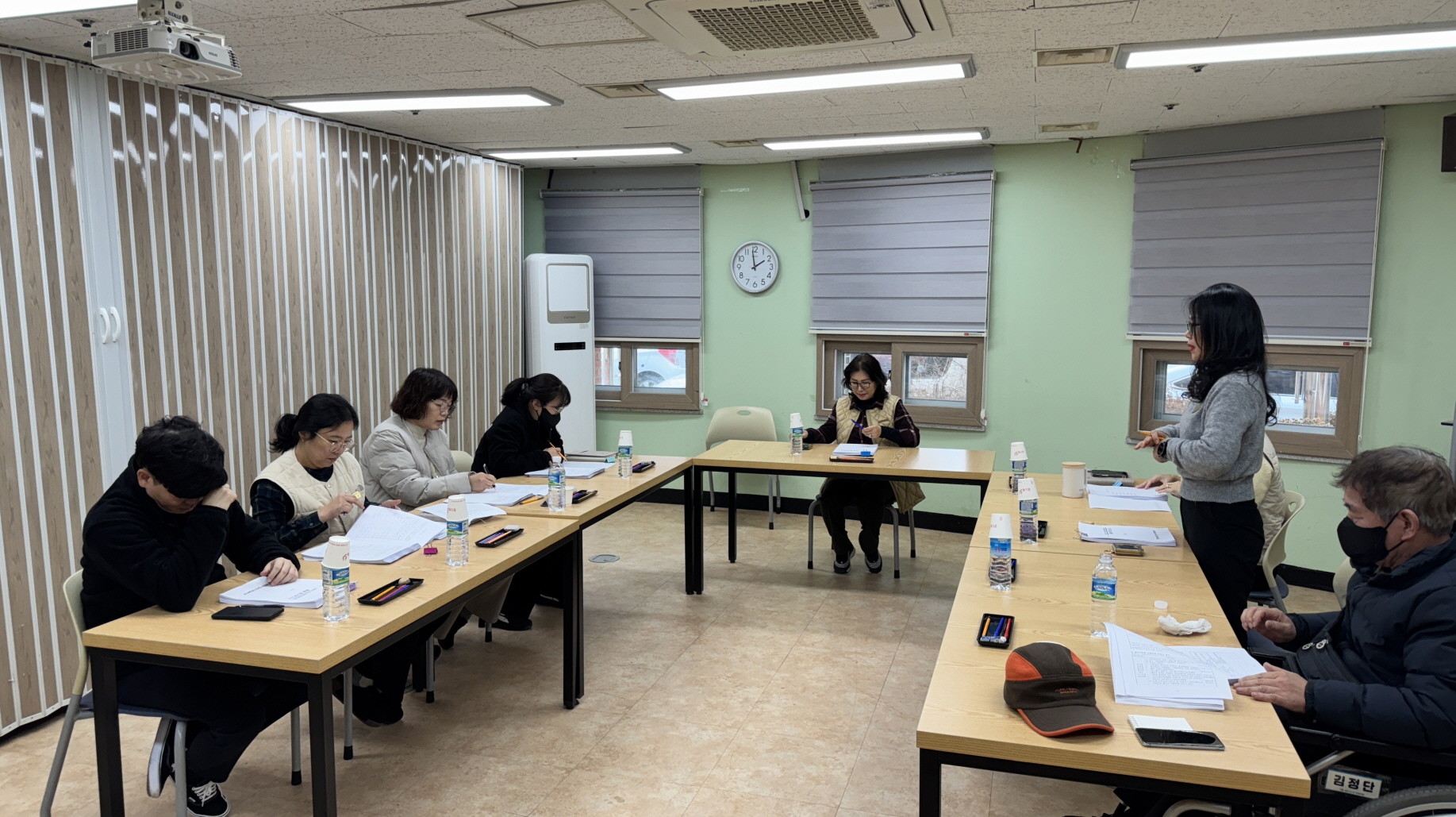 회의실에서 기획홍보팀장이 총괄 평가회의 자료를 발표하고 있으며, 다른 직원들은 책상에 앉아 문서를 검토하고 있다. 책상 위에는 물병과 자료가 놓여 있고, 뒤쪽에는 창문과 시계가 보인다.