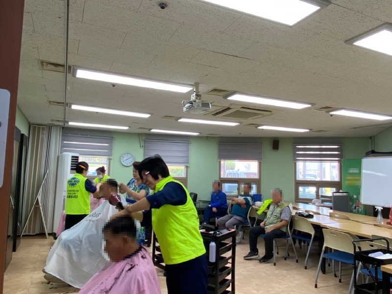 병아리봉사단 봉사자가 이용자의 머리를 다듬고 있고, 뒤편 의자에는 봉사를 기다리는 남성 이용자들이 앉아 있는 모습