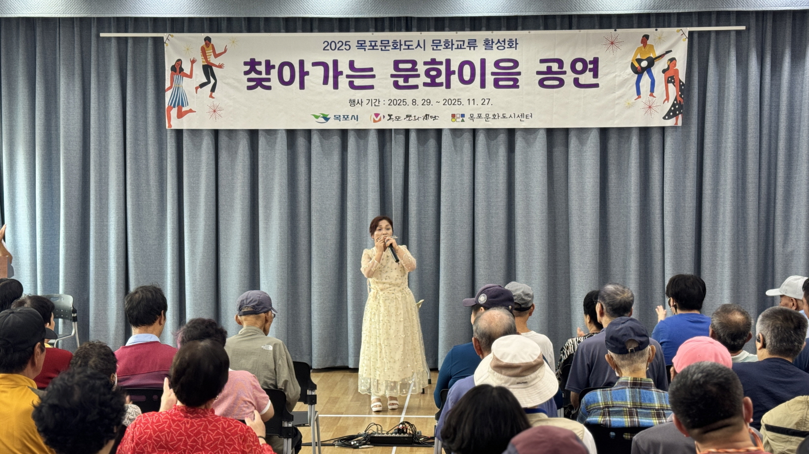연한 색 드레스를 입은 여성 가수가 무대 중앙에서 노래를 부르고 있으며, 관객들이 앞줄부터 가득 앉아 공연을 보고 있다. 가수 뒤로 회색 무대 커튼과 공연 현수막이 걸려 있다.