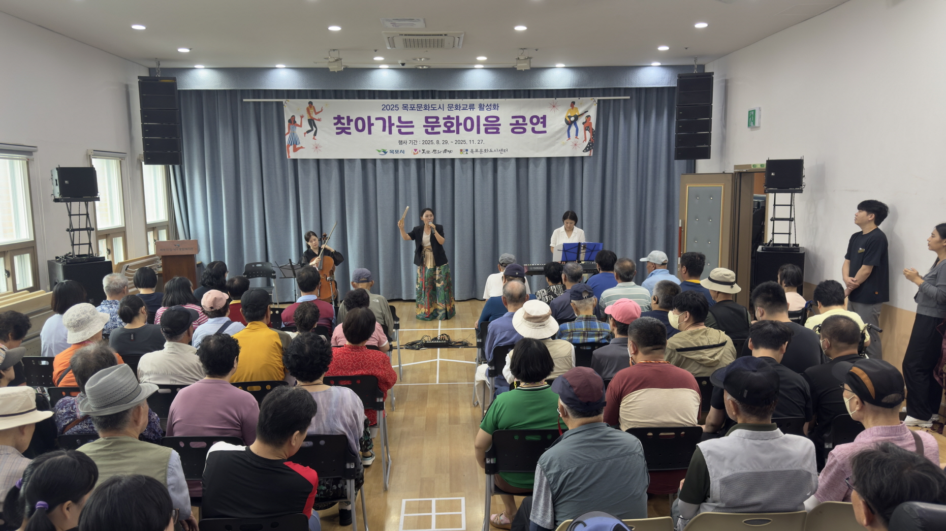 목포장애인종합복지관 강당 무대에서 ‘찾아가는 문화이음 공연’이 열리고 있다. 중앙에 가수와 첼로 연주자, 키보드 연주자가 함께 공연 중이며, 객석은 어르신과 장애인 관객들로 가득 차 집중해서 공연을 관람하고 있다.