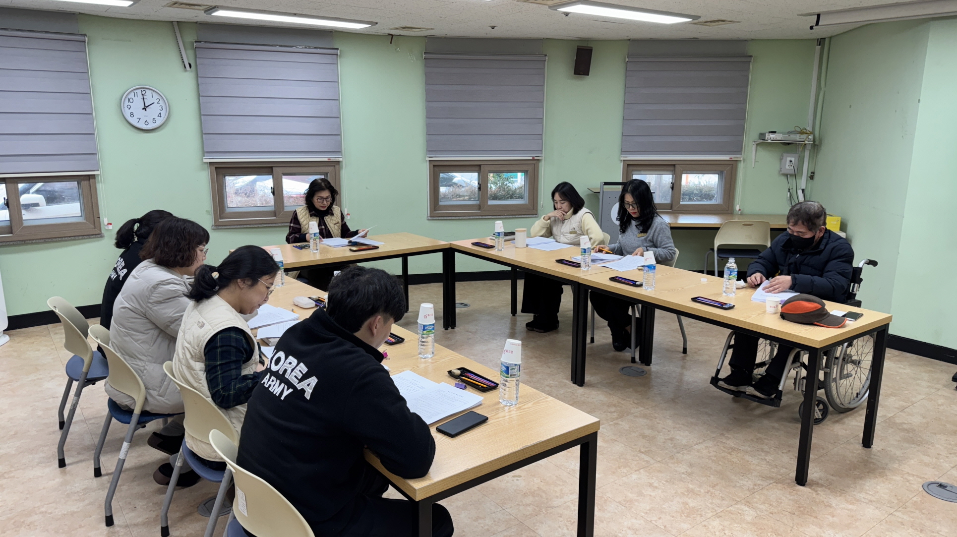 회의실에서 여러 명의 직원이 U자형으로 배치된 책상에 둘러앉아 문서를 보며 회의를 진행하고 있다. 책상 위에는 물병과 종이가 놓여 있으며, 오른쪽에는 휠체어를 이용하는 이용자협의회장이 앉아 회의에 참여하고 있다.