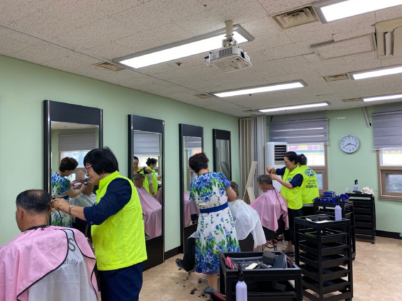 노란색 조끼를 입은 병아리봉사단 봉사자가 남성 이용자의 머리를 정성껏 깎고 있으며, 다른 봉사자들도 거울 앞에서 동시에 이·미용 봉사를 진행하는 모습