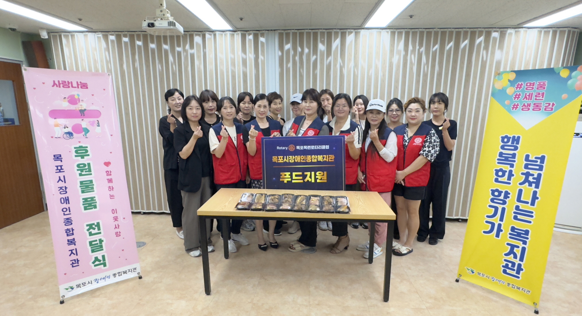 목련로터리클럽 회원들이 목포시장애인종합복지관 후원물품 전달식에서 푸드박스를 테이블에 올려놓고 함께 기념촬영을 하고 있다. 회원들이 손가락 하트를 하며 밝게 웃고 있으며, 양 옆에는 ‘후원물품 전달식’과 ‘행복한 복지관’ 현수막이 세워져 있다.