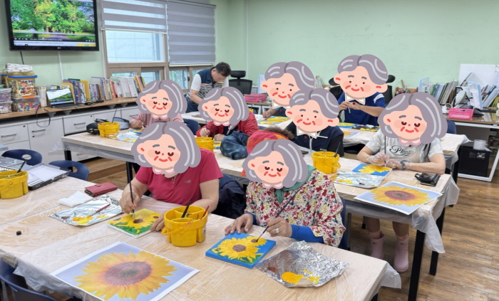여러 명의 어르신들이 복지관 미술실에서 해바라기 그림 그리기 활동에 참여하고 있다. 각자 캔버스와 붓, 물감을 사용해 해바라기 그림을 색칠하며 집중하는 모습이다.