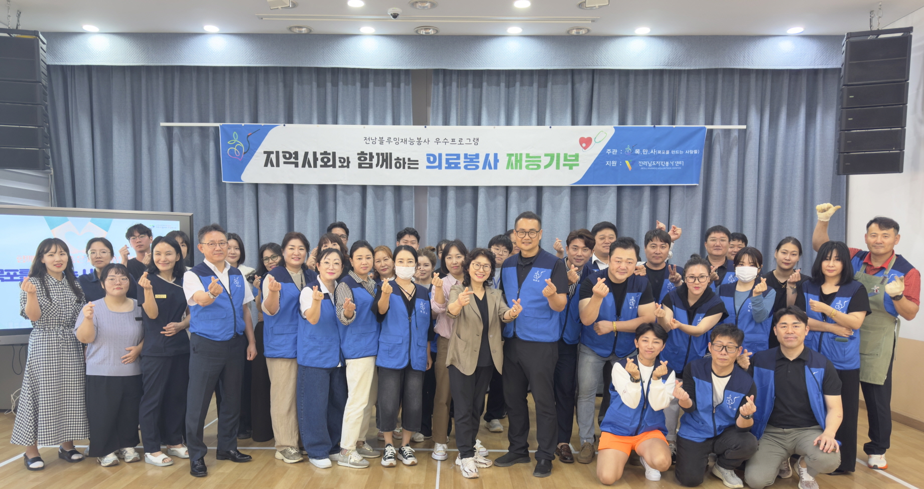목포를 만드는 사람들과 복지관 직원, 봉사자들이 함께 단체사진을 찍고 있다. 모두 파란색 조끼를 입고 손가락 하트를 하며 밝게 웃고 있다. 뒤에는 '지역사회와 함께하는 의료봉사 재능기부' 현수막이 걸려 있다.