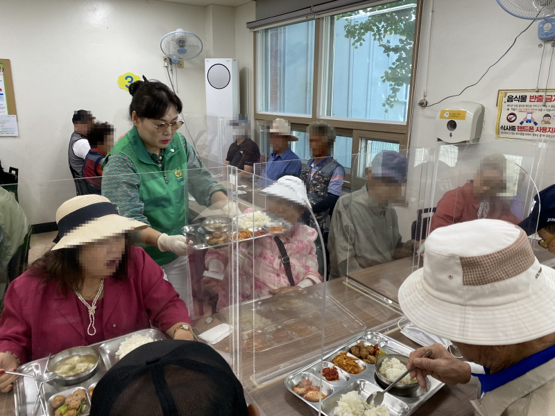 하당동새마을부녀회 봉사자가 초록색 조끼를 입고 점심 배식을 받아 앉아 있는 이용고객에게 음식을 전달하는 장면. 식탁 위에는 투명 가림막이 설치되어 있음