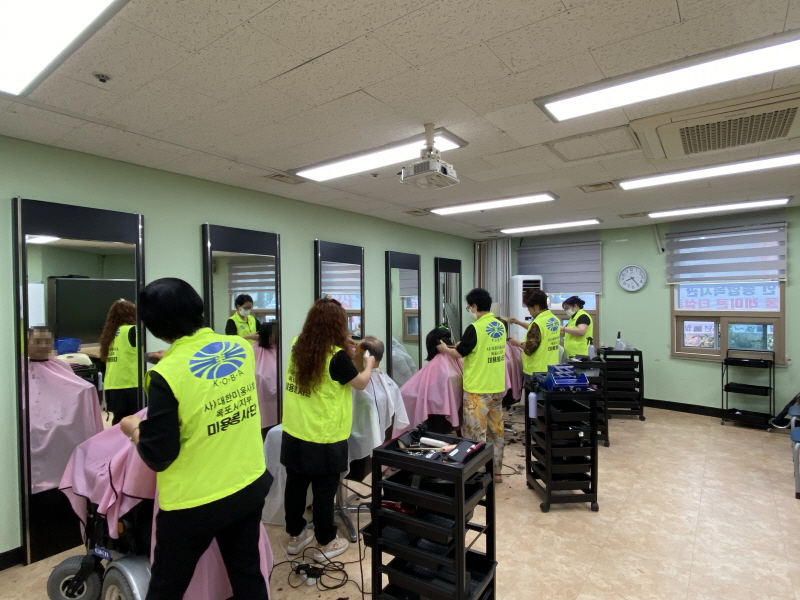 병아리봉사단 봉사자가 노란 조끼를 입고 이용고객들의 머리를 다듬고 있으며, 여러 명이 분홍색 커트를 두른 채 거울 앞에 앉아 있다