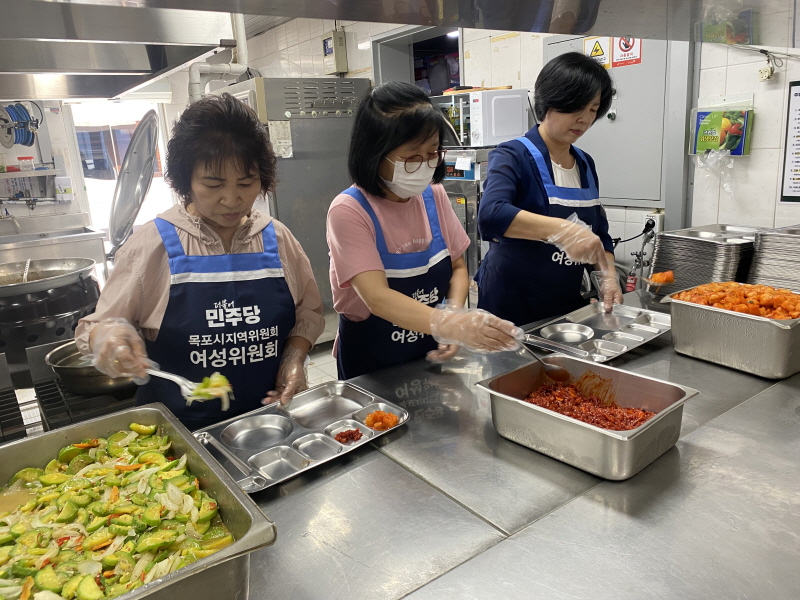 민주당여성위원회 봉사자들이 주방에서 반찬을 식판에 담는 모습. 테이블 위에는 애호박 볶음과 무침 반찬이 놓여 있음