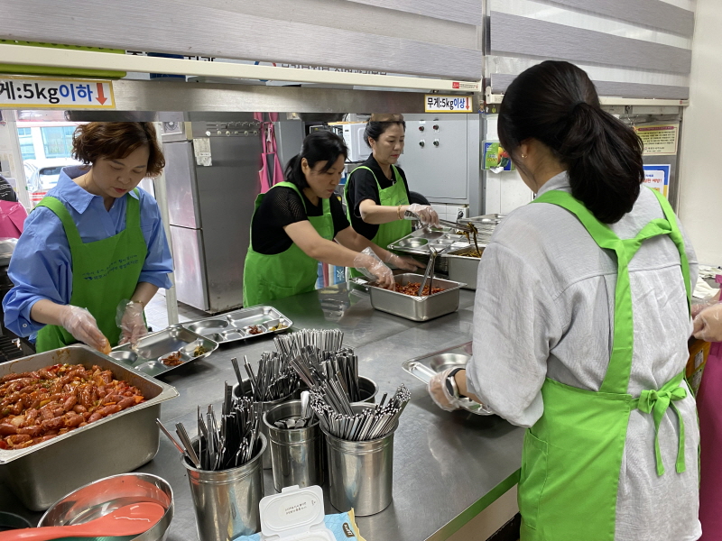 목포시청 공직자 부인회 봉사자들이 주방에서 김치와 반찬을 식판에 담고 있고, 앞에는 배식을 기다리는 사람이 서 있는 모습