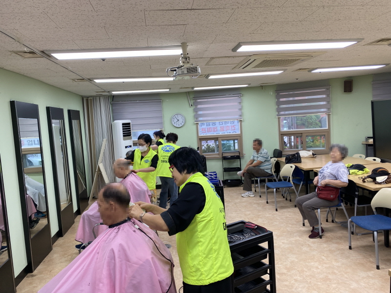 병아리봉사단 봉사자들이 커트를 두른 이용고객의 머리를 정성스럽게 다듬고 있고, 뒤쪽에는 차례를 기다리는 사람들이 앉아 있다