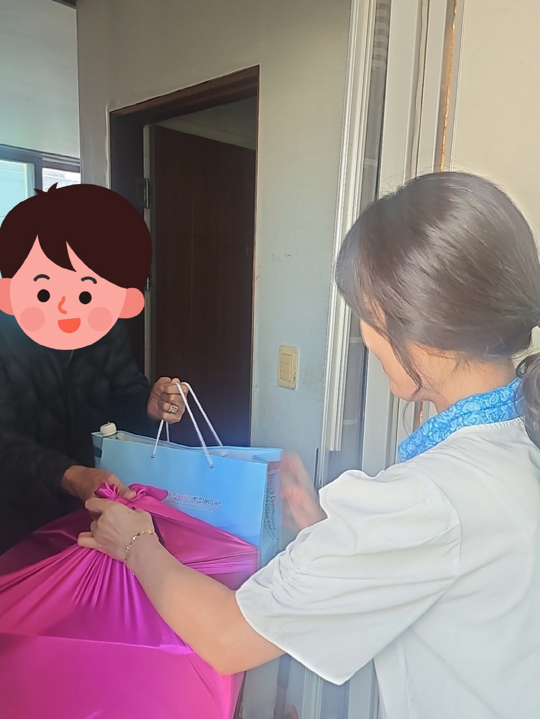 우리 복지관 직원이 문 앞에서 한 남성에게 분홍색 보자기와 선물 가방을 전해드리고 있다. 남성은 두 손으로 가방을 받으며 따뜻하게 맞이하고 있다