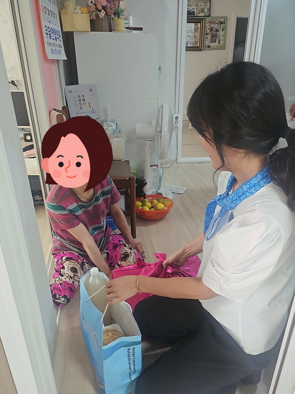 우리 복지관 직원이 무릎을 꿇고 앉아 분홍색 보자기와 선물 가방을 한 여성에게 전달하고 있다. 여성은 집 안에서 앉아 직원과 마주하며 감사한 표정을 짓고 있다