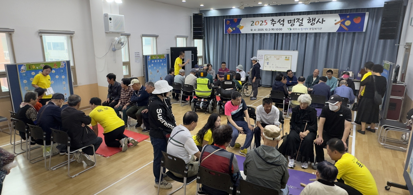 복지관 강당에서 열린 2025 추석 명절 행사 현장으로, 노란색 단체복을 입은 직원들이 여러 그룹으로 나뉘어 이용고객들과 윷놀이를 진행하고 있으며, 휠체어 이용고객도 함께 참여해 웃음과 대화가 오가는 활기찬 분위기다.