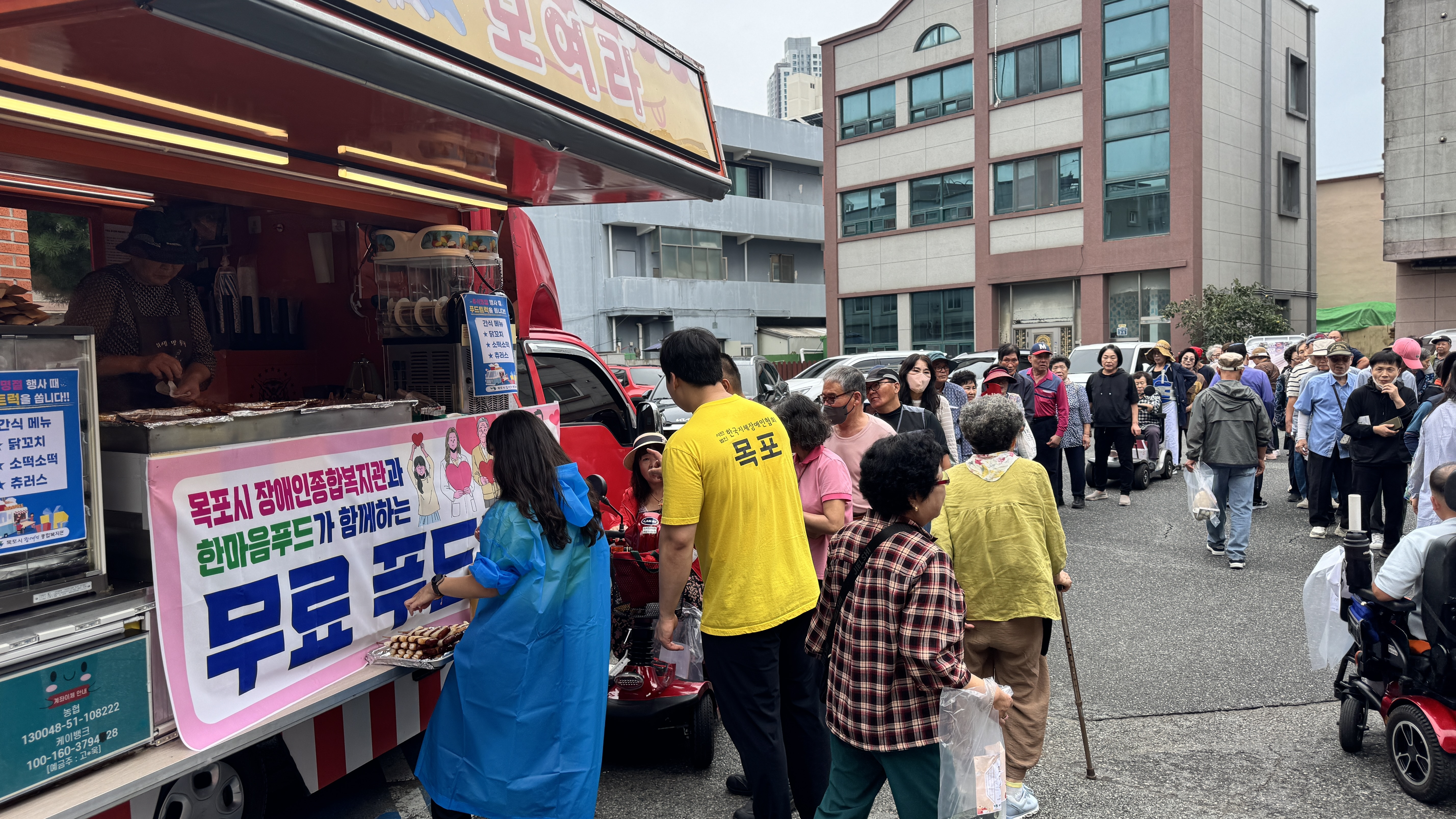 복지관 야외 주차장에서 열린 무료 푸드트럭 행사로, 붉은 푸드트럭 앞에서 직원과 이용고객들이 음식을 받아가며 줄을 서 있고, 뒤쪽으로 많은 사람들이 모여 활기찬 명절 분위기를 나누고 있다.