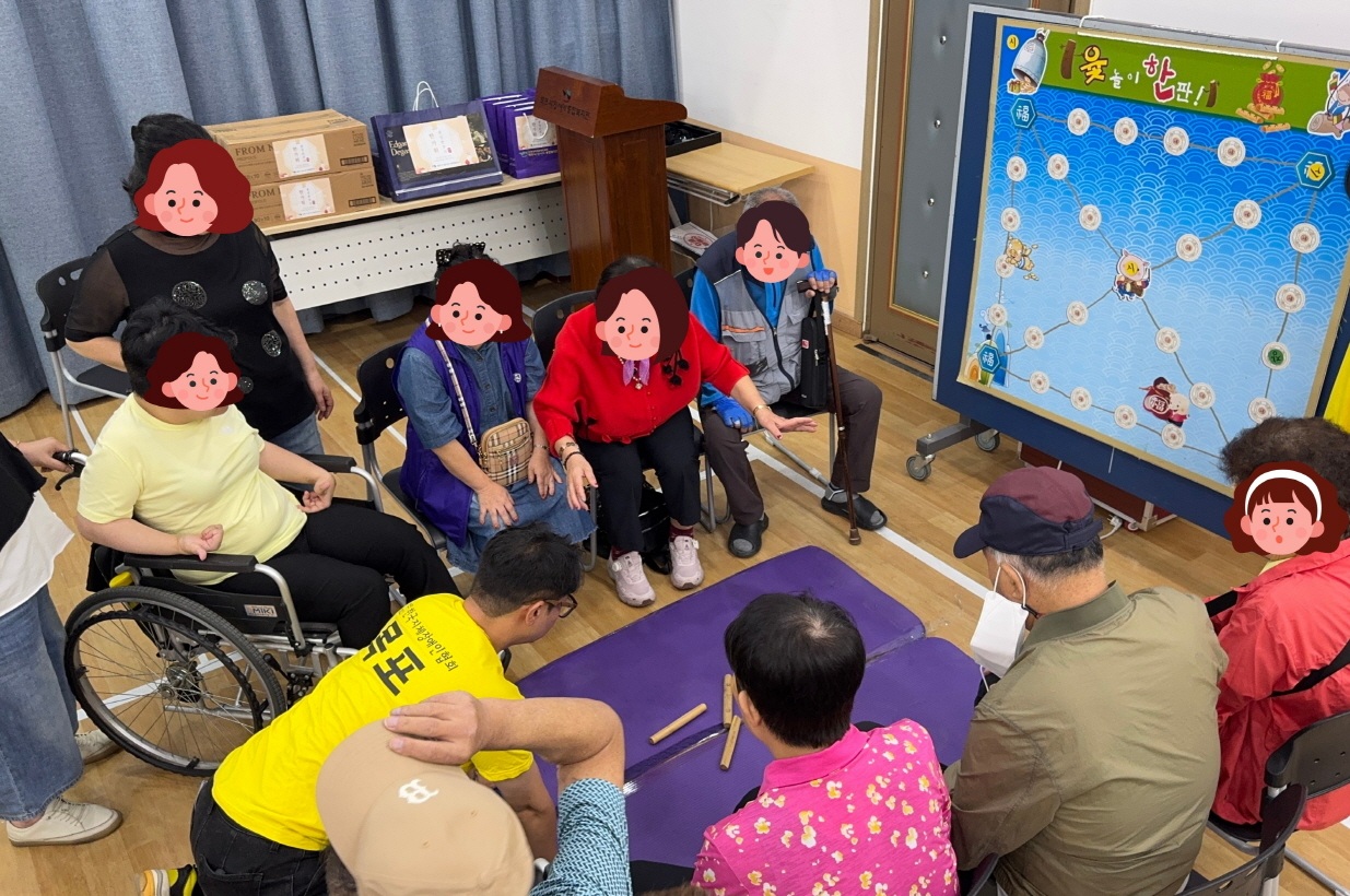 복지관 강당에서 이용고객이 바닥에 윷을 던지고 직원이 결과를 확인하며 웃고 있는 모습으로, 주변 이용고객들이 함께 응원하며 명절 분위기를 즐기고 있다.