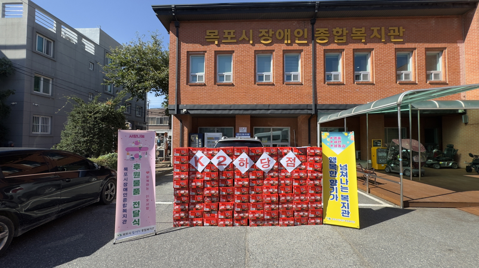 목포시장애인종합복지관 건물 앞마당에 ‘K2 하당점’에서 후원한 신라면 100박스가 높게 쌓여 있고, 좌우에는 ‘사랑나눔 후원물품 전달식’과 ‘행복이 넘쳐나는 복지관’이라고 적힌 배너가 세워져 있으며, 밝은 햇살 아래에서 따뜻한 나눔의 분위기가 느껴지는 장면이다.