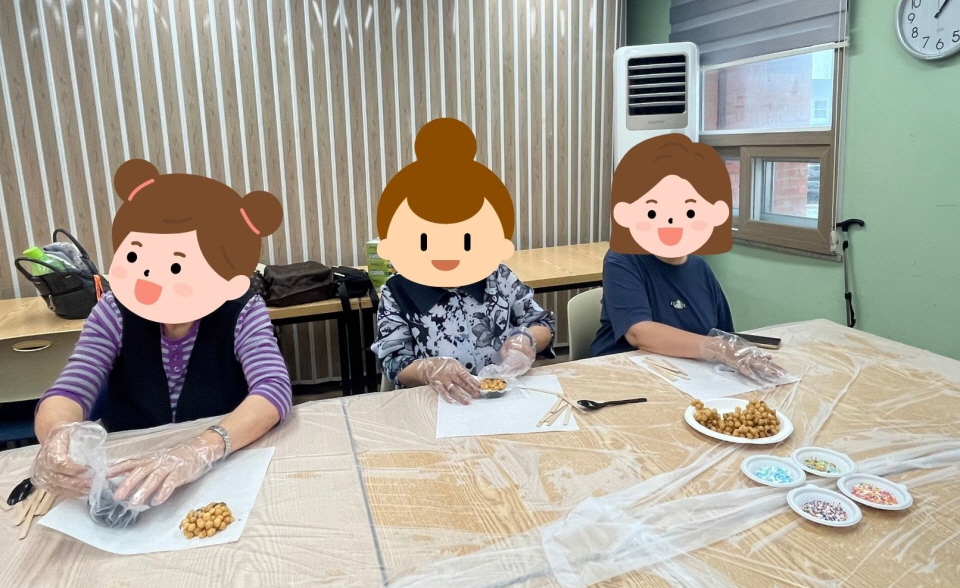 복지관 프로그램실에서 여성장애인들이 위생장갑을 끼고 전통 간식 만들기 활동을 하고 있음. 테이블 위에는 조청과자와 색색의 토핑 재료가 놓여 있고, 참여자들이 직접 손으로 모양을 빚으며 대화를 나누고 있음. 창가 쪽으로 밝은 햇살이 들어오는 따뜻한 분위기의 장면임.