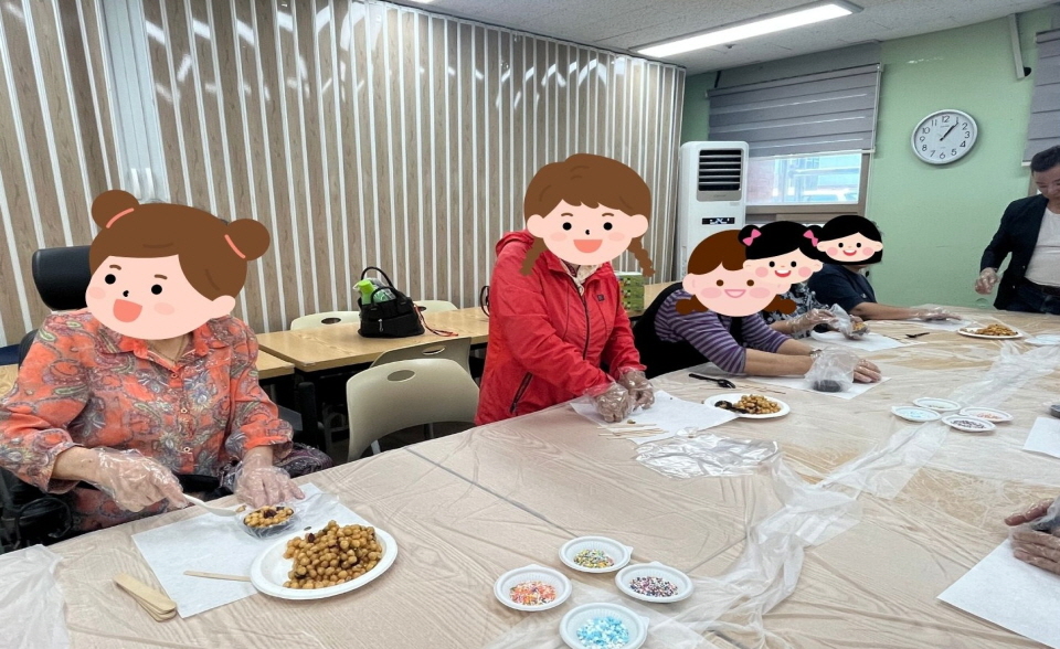 복지관 프로그램실에서 여성장애인들이 간식 만들기 활동에 참여하고 있음. 테이블 위에는 조청에 버무린 콩과자와 색색의 스프링클이 놓여 있으며, 참여자들이 위생장갑을 끼고 과자를 동그랗게 모양 내는 중임. 밝고 즐거운 분위기 속에서 서로 이야기를 나누며 손을 움직이고 있음.