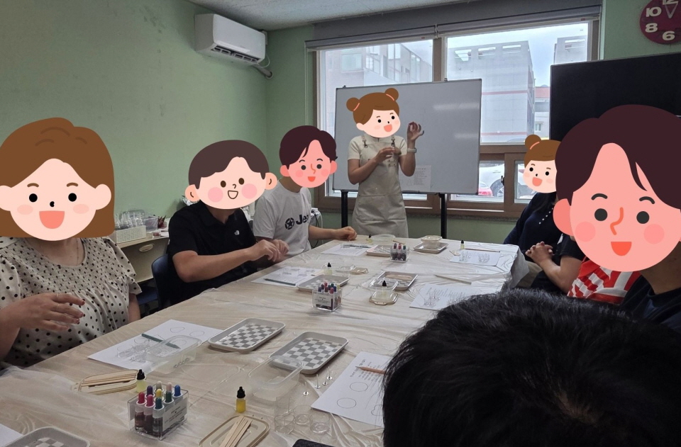 복지관 프로그램실에서 양초공예 체험이 이루어지고 있음. 강사가 앞에서 시연을 보이며 참여자들이 직접 재료를 섞고 향초를 완성하는 과정임.   테이블 위에 비커, 스포이드, 색소 등이 준비되어 있고, 참여자들이 서로 대화하며 즐겁게 활동하는 모습임.