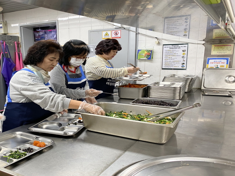 민주당여성위원회 봉사자 세 명이 복지관 조리실에서 배식 준비를 하고 있음. 파란색 앞치마를 착용하고 위생장갑을 낀 봉사자들이 반찬을 식판에 담고 있으며, 조리대 위에는 나물, 콩자반, 김치 등 여러 가지 반찬이 담긴 큰 통이 정돈되어 있음.