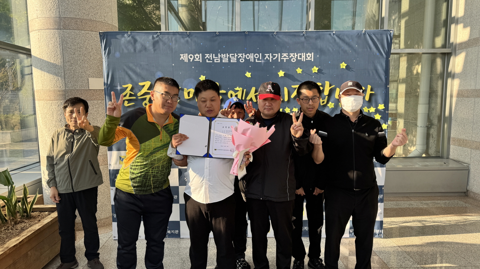 제9회 전남발달장애인 자기주장대회에서 목포시장애인종합복지관 참가자와 응원하러 온 이용고객들이 기념사진을 찍고 있음. 가운데 수상자가 도전상 상장과 꽃다발을 들고 있고, 주변의 이용고객들이 손가락 하트와 브이(V) 포즈를 하며 함께 축하하는 모습임. 뒤편에는 ‘존중은 마음에서 시작합니다’라는 문구가 적힌 행사 배너가 보임.