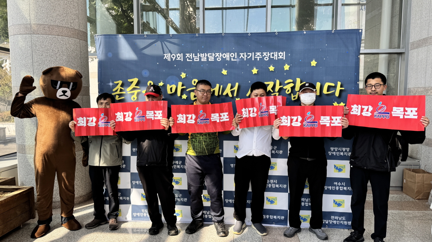 제9회 전남발달장애인 자기주장대회 행사장 입구에서 목포시장애인종합복지관 참가자와 응원하러 온 이용고객들이 단체로 기념 촬영을 하고 있음. 곰돌이 인형탈을 쓴 마스코트가 함께 서 있고, 이용고객들이 ‘최강 목포’라고 적힌 빨간색 응원 피켓을 들고 밝게 응원하는 모습임. 뒤편에는 ‘존중은 마음에서 시작합니다’라는 문구의 행사 배너가 걸려 있음.