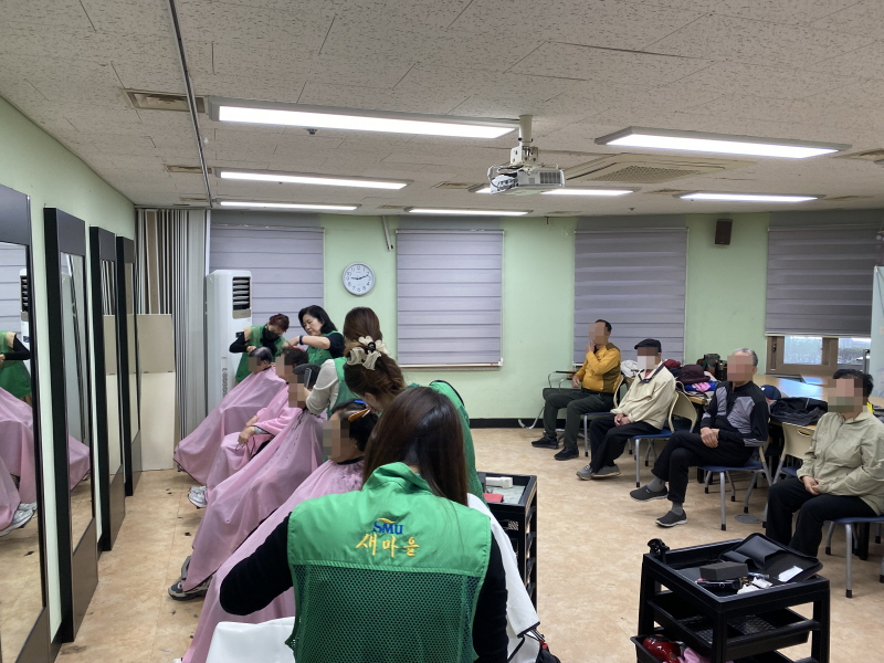 하당동새마을부녀회 봉사자들이 복지관 미용실에서 이미용 봉사를 진행하고 있음.   초록색 조끼를 입은 봉사자들이 이용고객의 머리를 손질하고 있으며, 대기석에는 여러 이용고객들이 차례를 기다리며 앉아 있음. 미용실 내부에는 거울과 의자가 나란히 배치되어 있음.