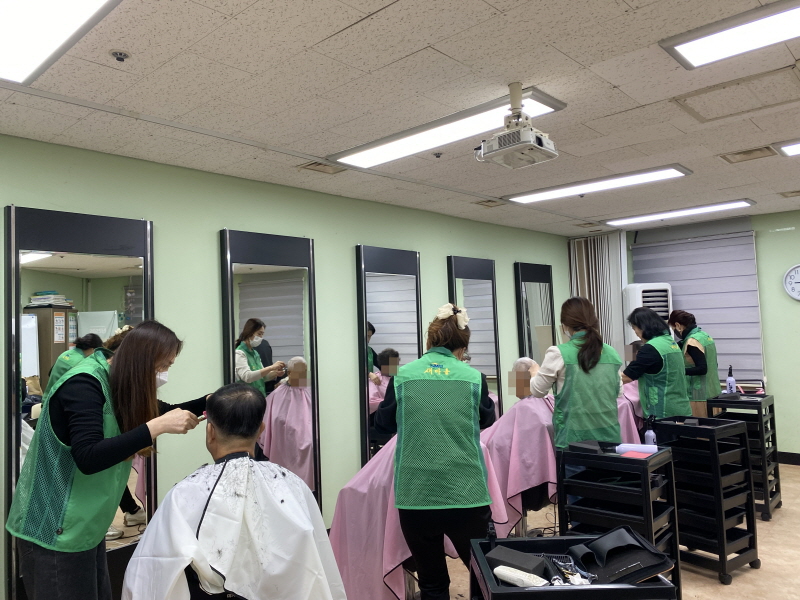 하당동새마을부녀회 봉사자들이 복지관 미용실에서 이용고객의 머리를 다듬고 있음. 초록색 조끼를 입은 봉사자들이 가위를 들고 머리를 자르고 있으며, 이용고객은 하얀색 가운을 두르고 있음. 거울 앞에는 미용 도구들이 정리되어 있고, 차분한 분위기 속에서 봉사가 진행되고 있음.