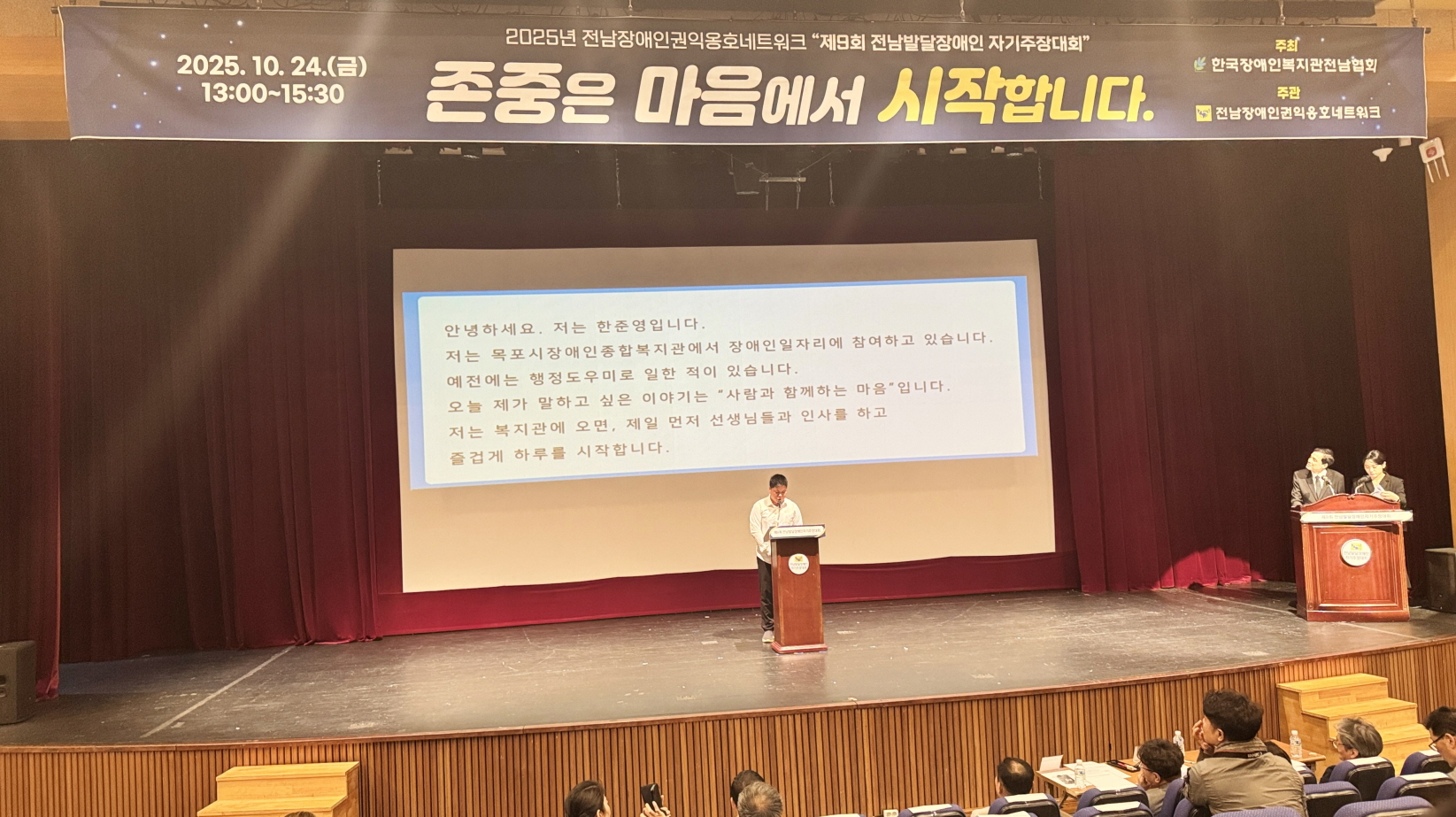 무대 위에서 목포시장애인종합복지관 이용고객이 연단에 서서 발표를 하고 있음. 대형 스크린에는 “안녕하세요. 저는 한준영입니다.”로 시작하는 발표문이 자막으로 표시되고, 무대 위 오른쪽에는 수어통역사가 함께 발표를 지원하고 있음. 상단 현수막에는 ‘2025년 전남장애인권익옹호네트워크 제9회 전남발달장애인 자기주장대회 – 존중은 마음에서 시작합니다’라고 적혀 있음.