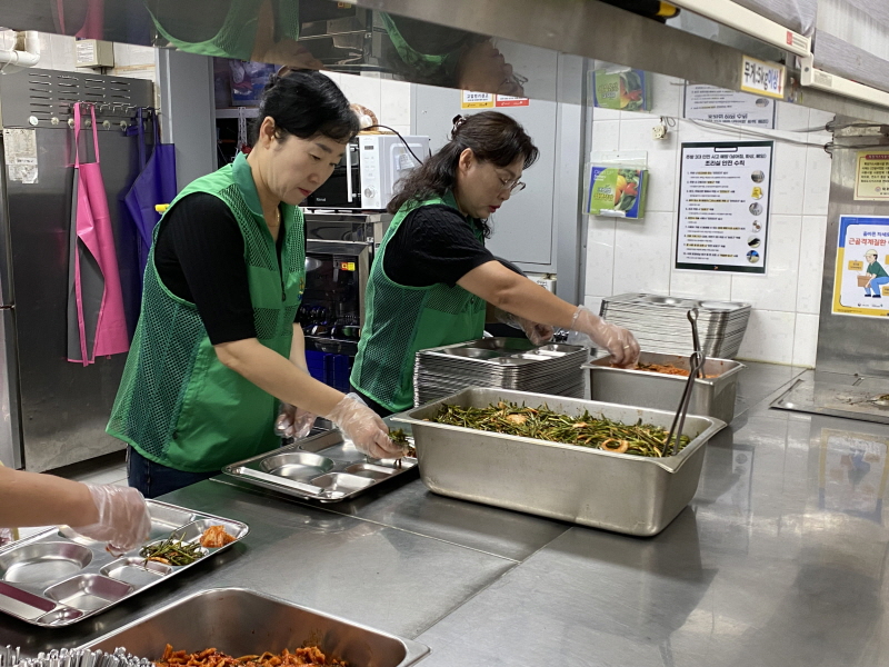 하당동새마을부녀회 봉사자 두 명이 복지관 조리실에서 배식 준비를 하고 있음. 초록색 조끼를 입은 봉사자들이 위생장갑을 착용하고 밥그릇에 나물을 담고 있으며, 조리대 위에는 여러 가지 반찬이 담긴 스테인리스 통이 놓여 있음.