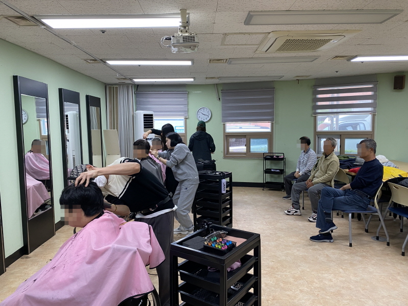 청담헤어 봉사자들이 복지관 미용실에서 이용고객의 머리를 정성껏 손질하고 있음. 검정색 복장과 편안한 옷차림의 봉사자들이 이용고객의 머리카락을 자르고 있으며, 창가 쪽 대기석에서는 다른 이용고객들이 차례를 기다리고 있음.