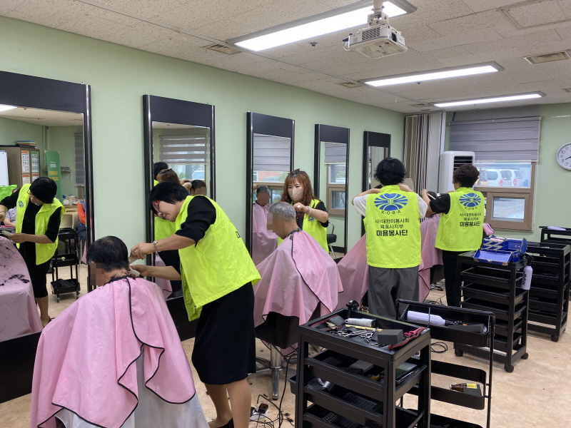 병아리봉사단 봉사자들이 복지관 미용실에서 이용고객의 머리를 다듬고 있음. 노란색 조끼를 입은 봉사자들이 머리카락을 자르고 있으며, 이용고객들은 분홍색 가운을 착용한 채 미용 의자에 앉아 있음. 거울과 미용도구가 가지런히 정리된 실내에서 단정한 분위기로 이미용 봉사가 진행되고 있음.