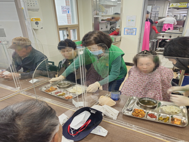 하당동새마을부녀회 봉사자가 식당 내에서 이용고객에게 식사를 전달하고 있음. 투명 칸막이 너머로 휠체어를 이용하는 고객에게 밥과 국, 반찬이 담긴 식판을 건네며 도움을 주는 모습. 다른 이용고객들도 식탁에 앉아 식사 준비를 하고 있음.
