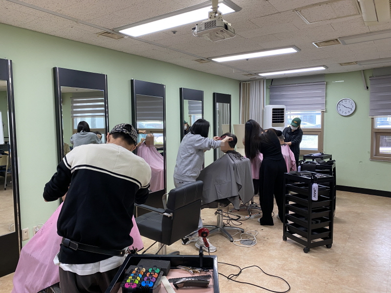 청담헤어 봉사자들이 여러 명의 이용고객을 대상으로 이미용 봉사를 진행하고 있음. 거울 앞 의자마다 고객이 앉아 머리를 자르고 있으며, 봉사자들은 드라이기와 미용기구를 사용해 머리를 정리함. 실내는 따뜻하고 밝은 조명 아래 깔끔하게 정돈된 모습임.