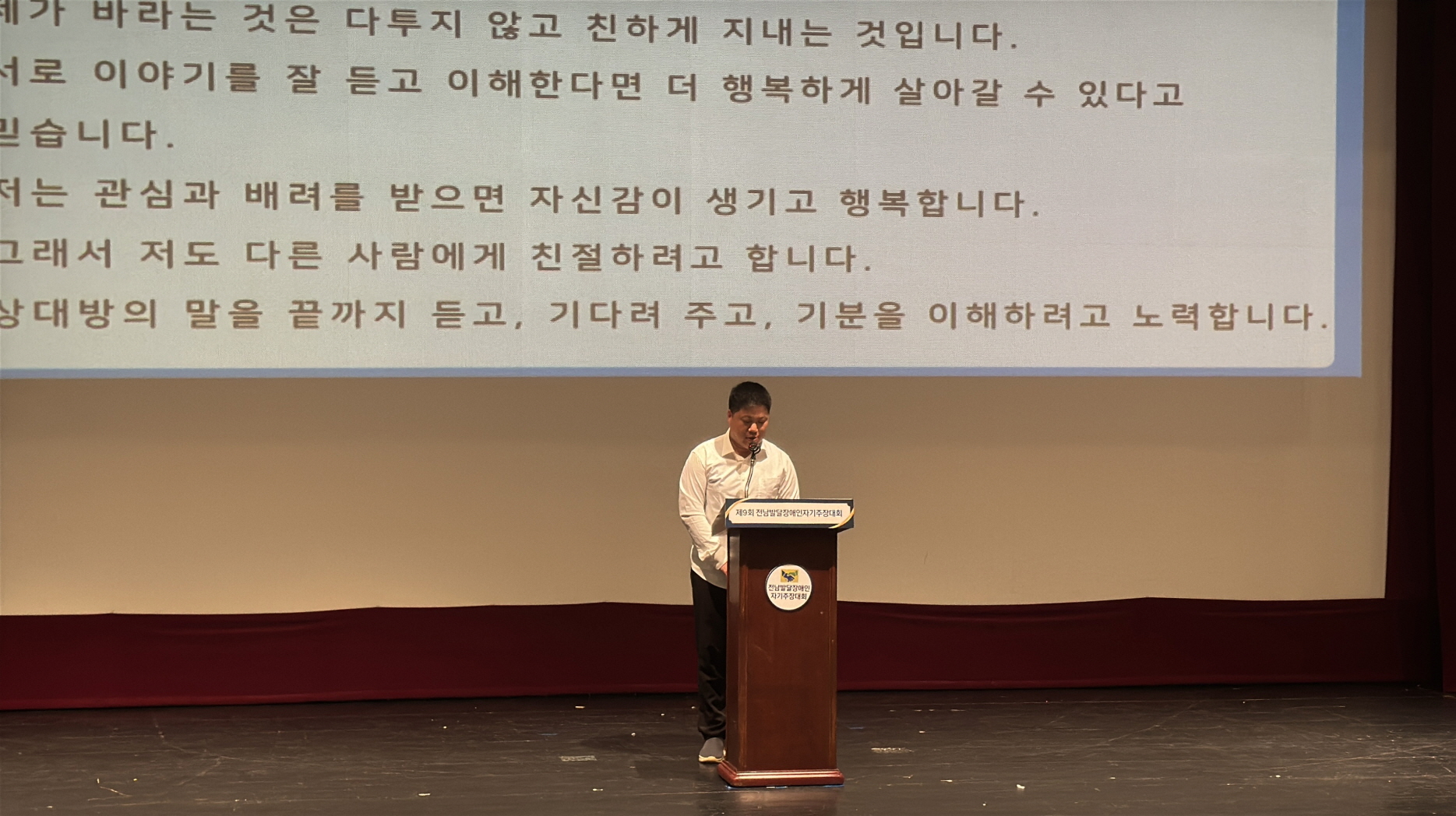 무대 중앙에서 발표자가 연단에 서서 발표를 이어가고 있음. 배경 스크린에는 “저는 관심과 배려를 받으면 자신감이 생기고 행복합니다.”라는 문장이 보임. 청중석에는 관람객들이 조용히 발표를 경청하고 있음.