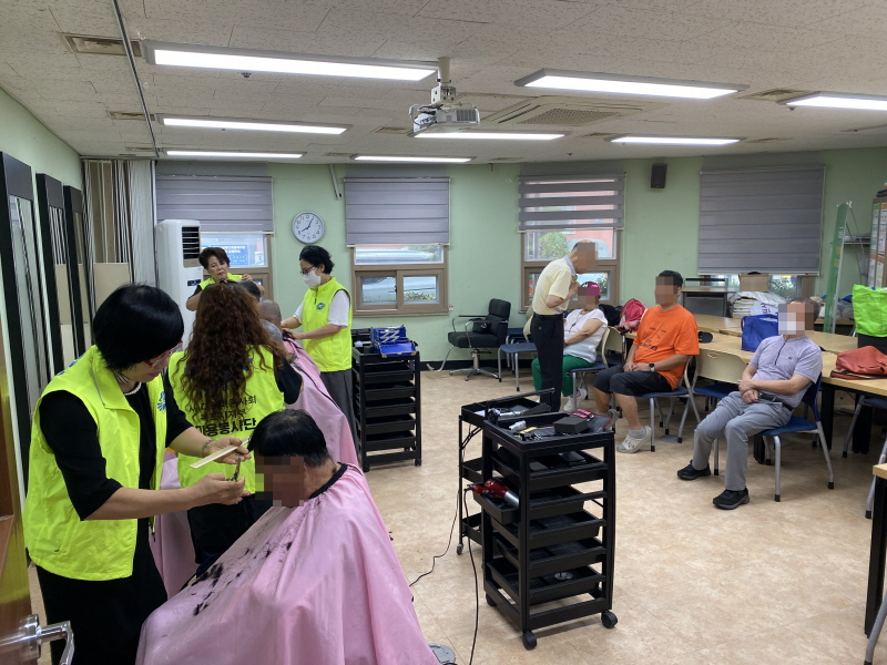 병아리봉사단 봉사자들이 이용고객의 머리를 손질하고 있으며, 다른 이용고객들은 대기석에서 순서를 기다리고 있음. 노란색 조끼를 입은 봉사자들이 세심하게 머리를 다듬는 모습이며, 복지관 미용실 안에는 차분한 분위기 속에서 대기 중인 고객들이 편히 앉아 있음.