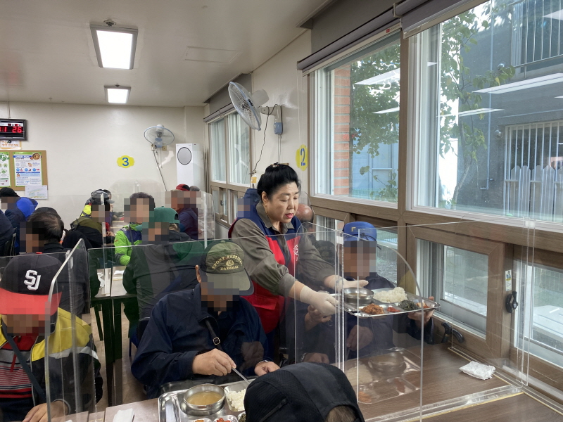 목련로터리클럽 봉사자가 복지관 식당에서 이용고객에게 식판을 전달하고 있음. 빨간 조끼를 입은 봉사자가 창가 쪽 테이블에 앉은 이용고객에게 밥과 반찬이 담긴 식판을 건네는 모습. 식당 내부에는 투명 칸막이가 설치되어 있고, 여러 이용고객이 식사 중임.
