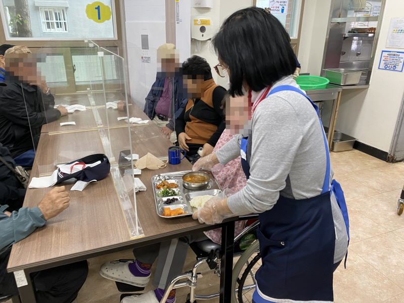 민주당여성위원회 봉사자가 복지관 식당에서 이용고객에게 식판을 전달하고 있음. 마스크를 착용한 봉사자가 따뜻한 국과 밥, 반찬이 담긴 식판을 휠체어 이용고객에게 건네며 식사를 돕고 있음. 주변에는 식사 중인 다른 이용고객들이 자리하고 있음.