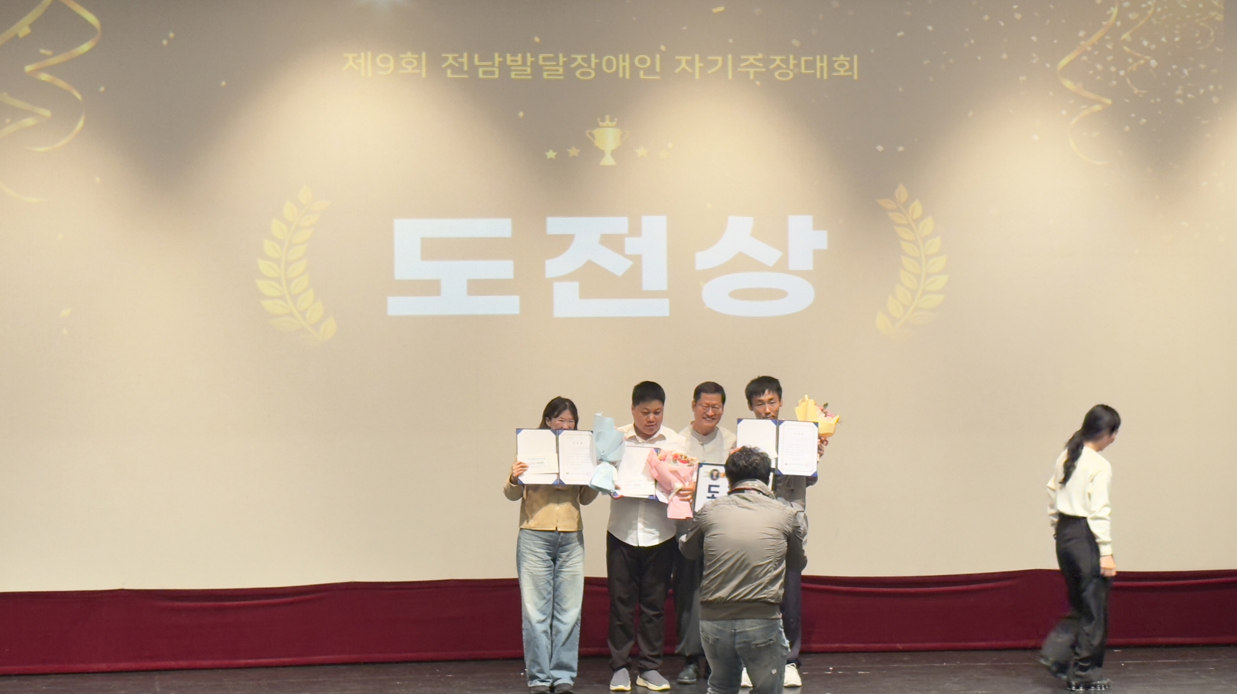 ‘제9회 전남발달장애인 자기주장대회’ 시상식 장면. 무대 스크린에 ‘도전상’이라는 글자가 크게 비춰지고 있으며, 수상자인 이용고객이 다른 참가자들과 함께 상장과 꽃다발을 들고 기념사진을 찍고 있음. 앞에서는 사진작가가 수상 장면을 촬영 중임.