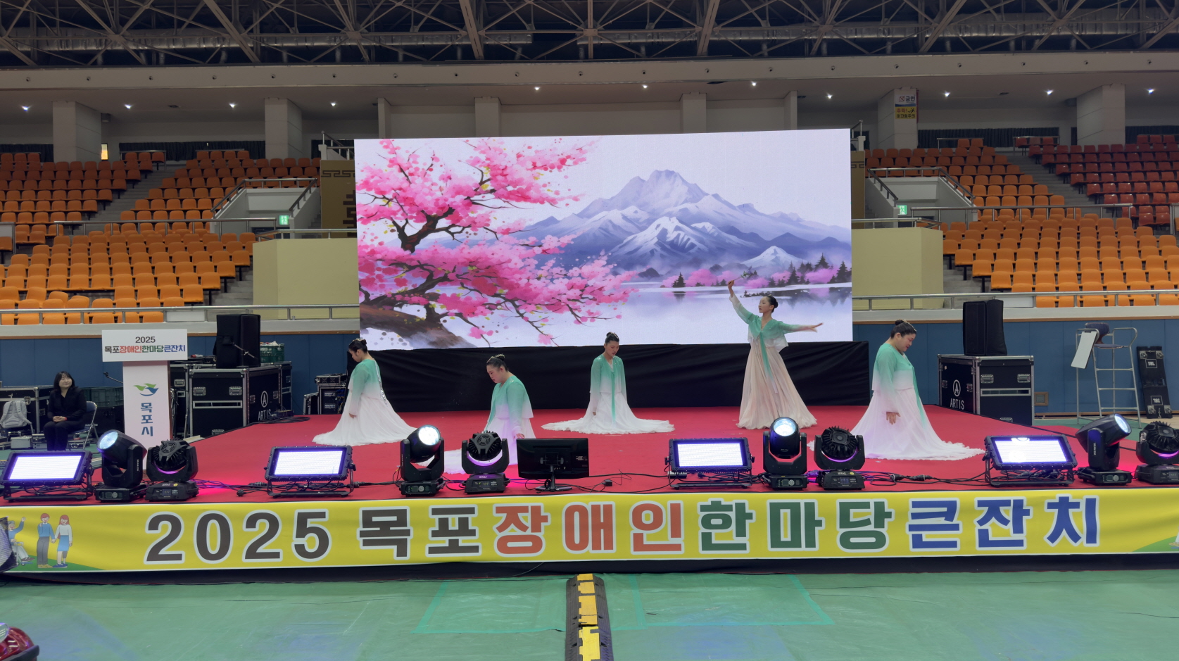 실내 체육관 무대 위에서 공생재활원 이용고객들이 민트색과 흰색이 그라데이션 된 긴 치마를 입고 기관공연으로 단체 무용을 선보이고 있으며, 뒤 대형 전광판에는 벚꽃과 설산 풍경 그림이 펼쳐져 있는 모습.